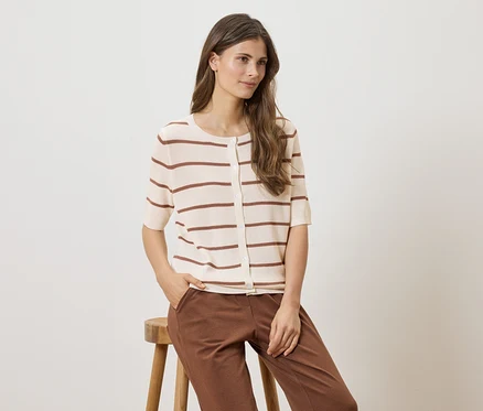 Une femme assise sur un tabouret, portant un cardigan rayé à manches courtes et un pantalon marron.