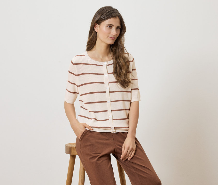Une femme assise sur un tabouret, portant un cardigan rayé à manches courtes et un pantalon marron.