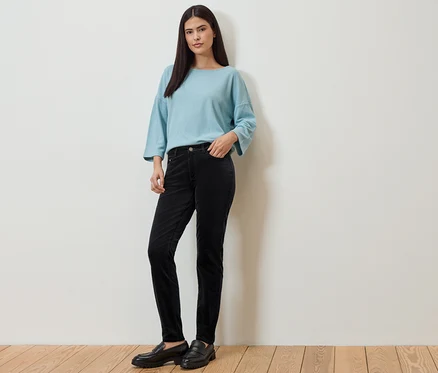 Une femme portant un haut bleu clair et un pantalon en velours côtelé noir pose devant un mur blanc. Elle porte des mocassins noirs.