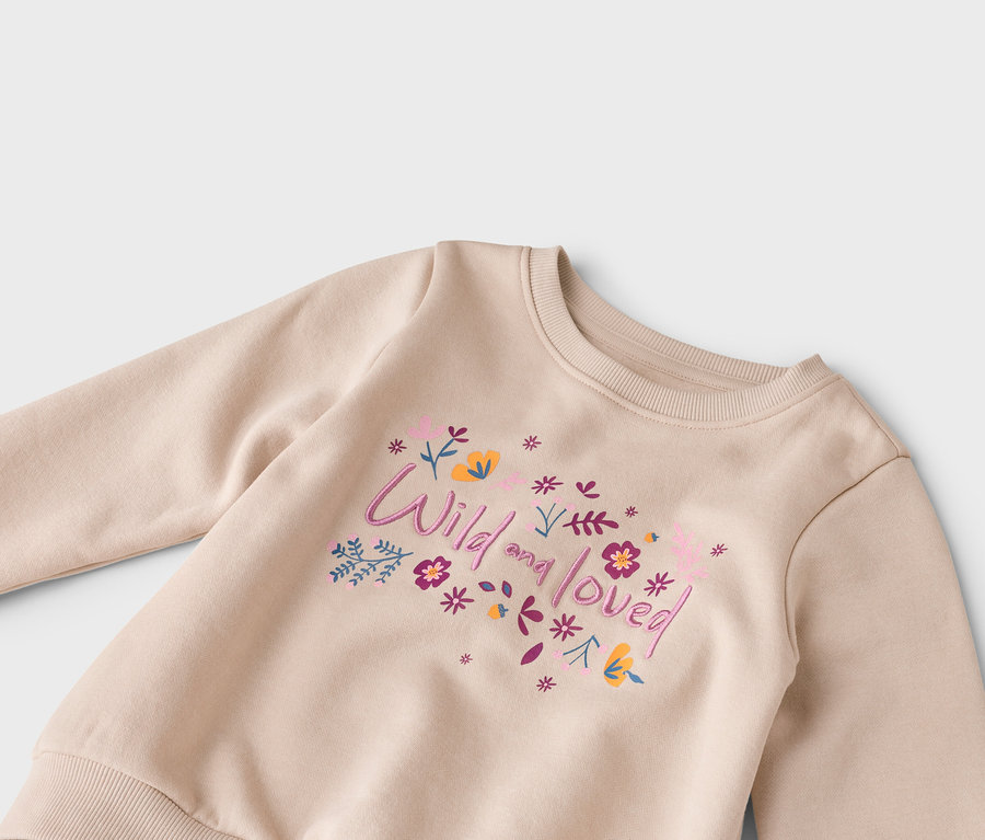 Gros plan d'un sweat-shirt beige avec l'inscription « Wild and loved » et des fleurs roses, violettes et jaunes.