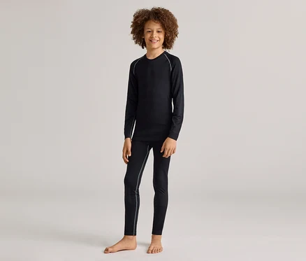 Un jeune garçon souriant aux cheveux bouclés se tient pieds nus, vêtu d'un haut noir à manches longues et d'un legging noir assorti avec des coutures gris clair.