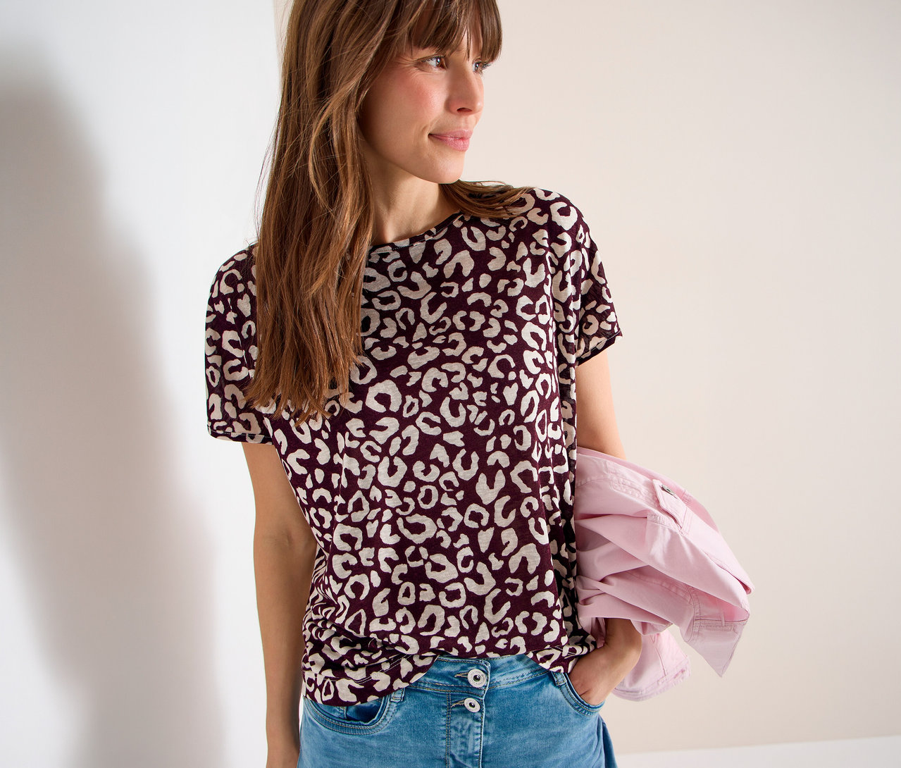Femme portant un t-shirt bordeaux à motif léopard et un jean, une veste rose sur le bras.