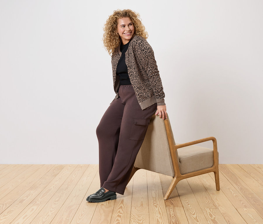 Une femme aux cheveux bouclés s'appuie contre un fauteuil, portant un pantalon marron, un pull noir et un cardigan tricoté marron et noir à motif animal.