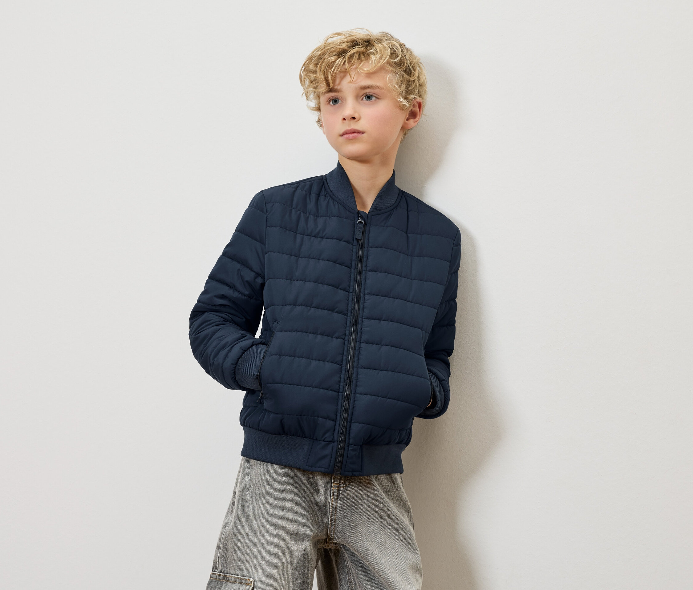 Un garçon blond portant un blouson matelassé bleu marine et un jean gris pose devant un mur blanc.