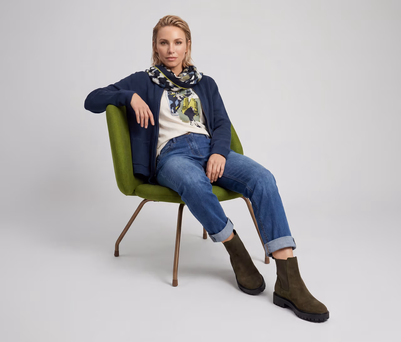 Une femme blonde est assise dans un fauteuil vert, vêtue d'une veste bleu marine, d'un t-shirt crème à motif, d'un jean bleu retroussé, d'une écharpe à motifs et de bottes vert olive.