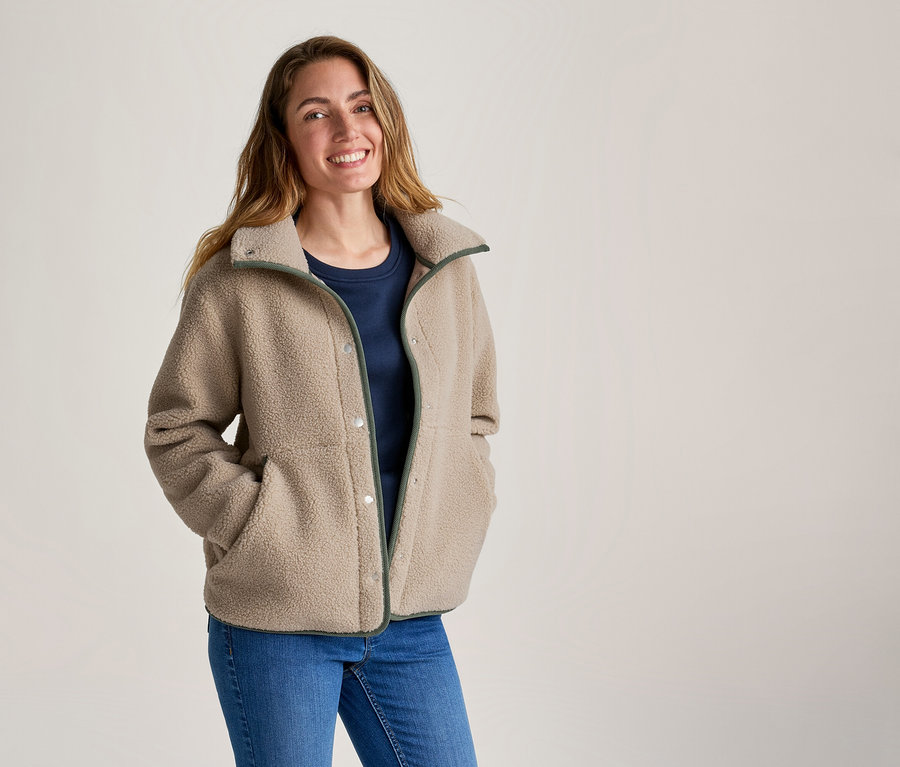 Femme souriante portant une veste polaire beige à bordure verte, un t-shirt bleu marine et un jean, les mains dans les poches de la veste.