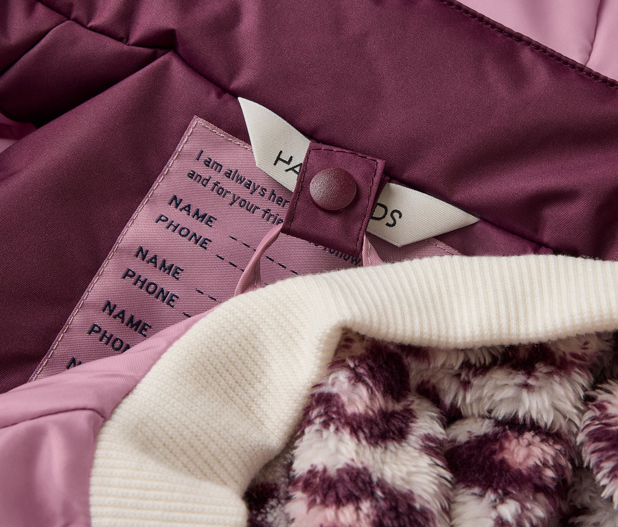 Gros plan de la doublure intérieure d'une veste violette avec un poignet côtelé blanc et une doublure moelleuse à motifs. Une étiquette avec des champs pour le nom et le numéro de téléphone est visible.
