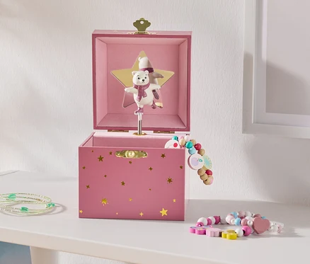 Une boîte à musique rose ouverte avec des étoiles dorées et un ours polaire dansant sur une étoile dorée, à côté de bracelets de perles colorés sur une étagère blanche.