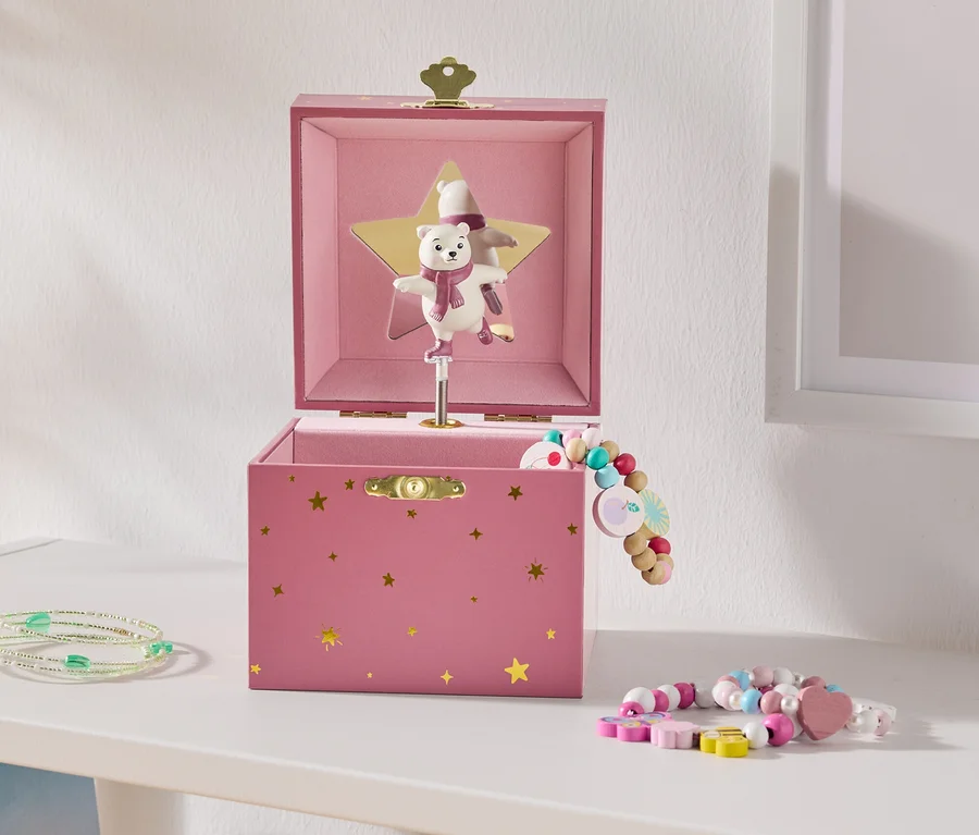 Une boîte à musique rose ouverte avec des étoiles dorées et un ours polaire dansant sur une étoile dorée, à côté de bracelets de perles colorés sur une étagère blanche.