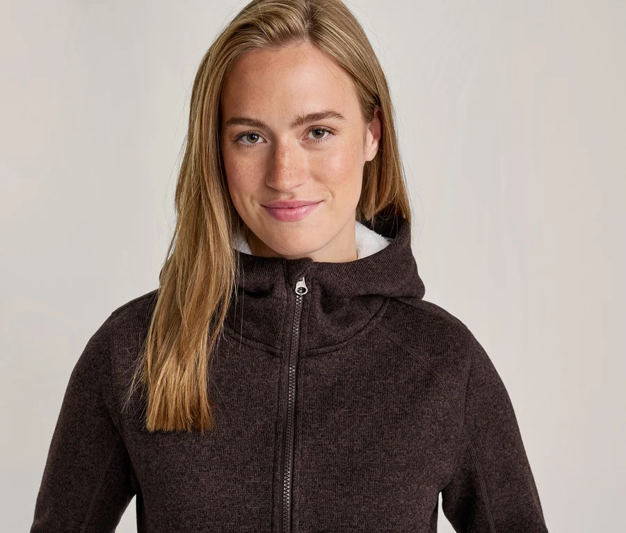 Femme souriante aux longs cheveux blonds portant une veste à capuche zippée marron foncé texturée avec une doublure blanche duveteuse.