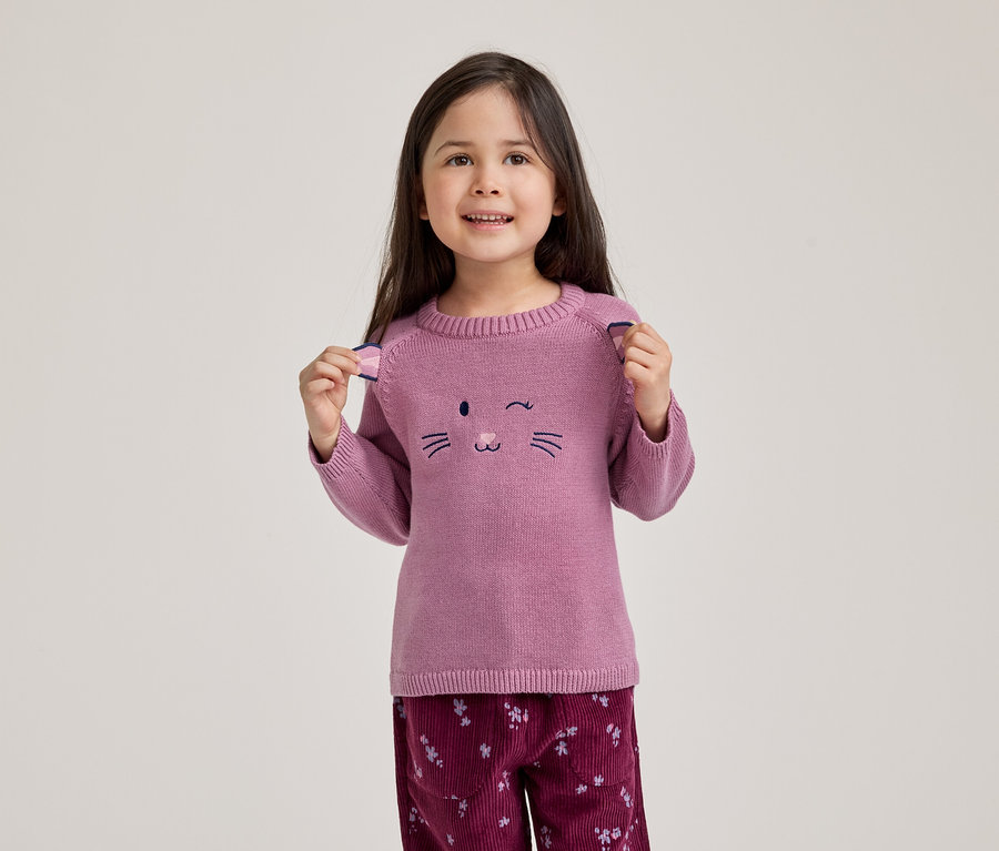 Une jeune fille souriante aux cheveux foncés porte un pull rose avec un motif de visage de chat clignotant et un pantalon en velours côtelé floral rouge foncé. Elle tient les petits détails en forme d'oreilles sur les épaules de son pull.