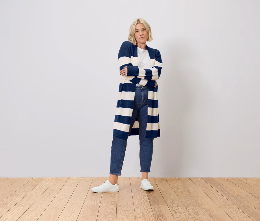 Femme blonde aux bras croisés, portant un long cardigan rayé bleu marine et blanc, un jean et des baskets blanches.