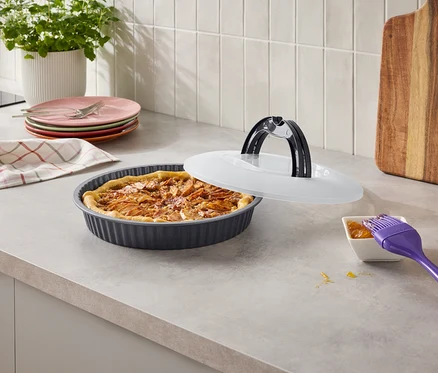 Une tarte aux fruits cuite dans un moule cannelé gris foncé repose sur un comptoir de cuisine, partiellement recouverte d'un couvercle blanc. À proximité se trouvent des assiettes colorées, un torchon à carreaux, une plante en pot, une planche à découper en bois et un petit bol de confiture avec un pinceau violet.
