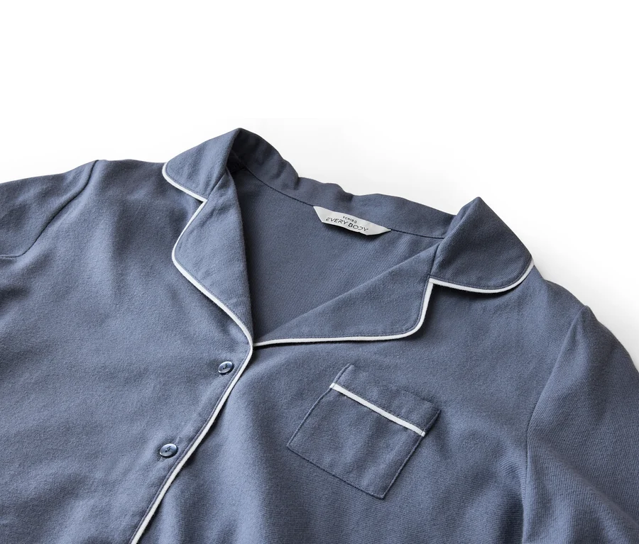 Gros plan d'un haut de pyjama bleu-gris avec passepoil blanc sur le col, la patte de boutonnage et la poche poitrine.