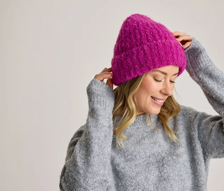 Femme souriante en pull gris ajustant un bonnet tricoté rose.