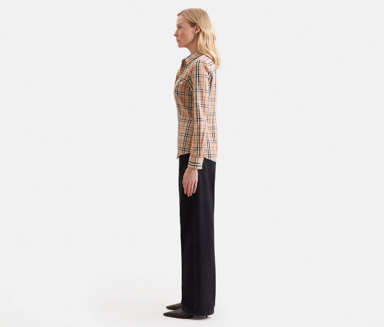 Femme blonde de profil, portant une chemise à manches longues à carreaux beige et un pantalon large foncé.