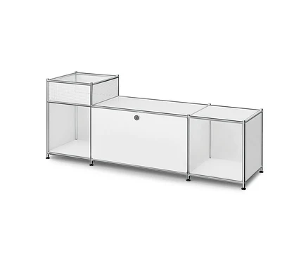 Meuble modulaire blanc avec cadre chromé. Il comporte en haut à gauche un compartiment ouvert aux côtés perforés, en dessous une étagère ouverte, au centre un meuble fermé avec une poignée ronde et à droite une autre étagère ouverte. L'unité repose sur de petits pieds noirs.