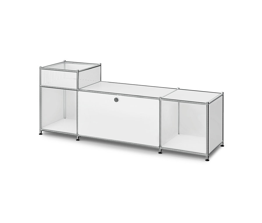 Meuble modulaire blanc avec cadre chromé. Il comporte en haut à gauche un compartiment ouvert aux côtés perforés, en dessous une étagère ouverte, au centre un meuble fermé avec une poignée ronde et à droite une autre étagère ouverte. L'unité repose sur de petits pieds noirs.