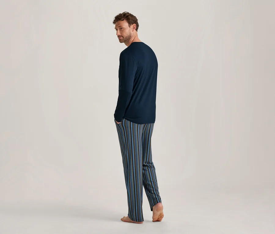 Homme vu de dos, regardant par-dessus son épaule gauche, portant un t-shirt à manches longues bleu marine et un pantalon de pyjama rayé bleu, marron et bleu clair, pieds nus.