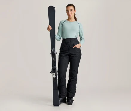 Une femme se tient debout, tenant un ski noir à la verticale, vêtue d'un haut à manches longues vert clair et d'un pantalon de ski noir taille haute.