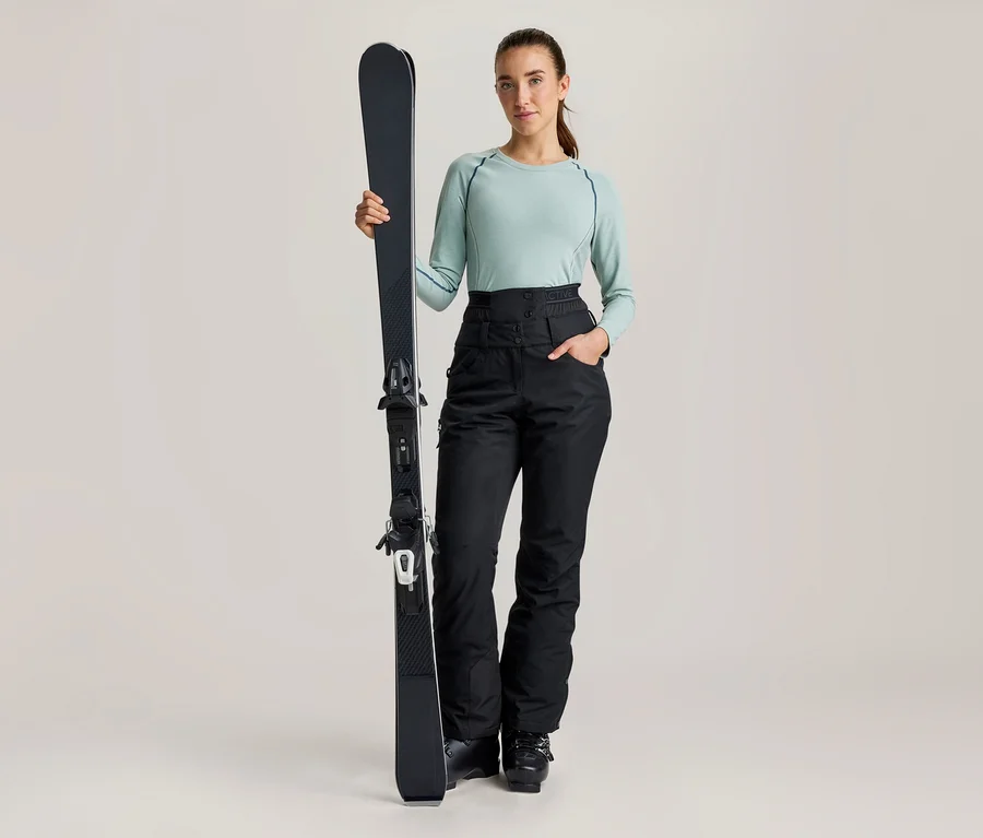 Une femme se tient debout, tenant un ski noir à la verticale, vêtue d'un haut à manches longues vert clair et d'un pantalon de ski noir taille haute.