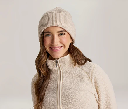 Femme souriante aux longs cheveux bruns portant un bonnet côtelé beige et une veste polaire crème zippée.