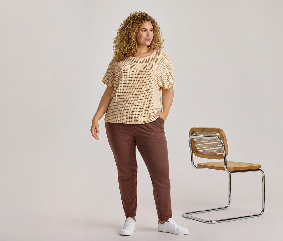 Une femme aux cheveux blonds bouclés, vêtue d'un haut rayé clair, d'un pantalon marron foncé et de baskets blanches, se tient à côté d'une chaise en bois moderne.