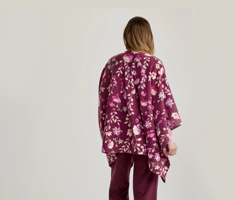 Une personne vue de dos, portant un poncho floral bordeaux avec des fleurs roses, violettes et blanches et un pantalon bordeaux assorti, sur un fond clair uni.
