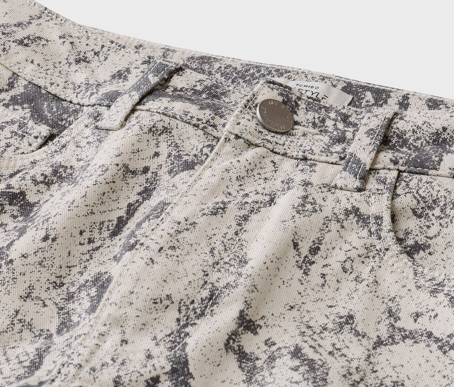 Gros plan sur la taille d'un pantalon beige clair à motif moucheté gris foncé, avec un bouton argenté, des passants de ceinture et une étiquette « TCHIBO STYLE ».