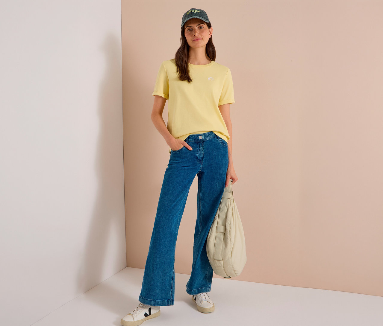 Femme portant un t-shirt jaune, un jean large bleu, des baskets blanches et une Casquette de baseball CECIL vert foncé, tenant un sac rembourré de couleur claire.
