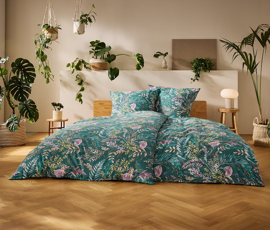 Un lit avec une parure de lit en renforcé verte à motifs floraux, dans une chambre lumineuse avec parquet et de nombreuses plantes en pot.