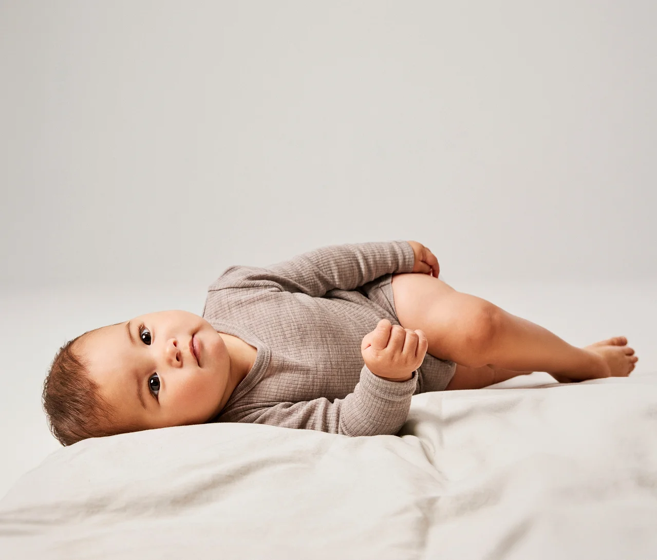 Bébé allongé sur le dos sur un drap clair, portant un body côtelé à manches longues de couleur taupe, regardant l'appareil photo.