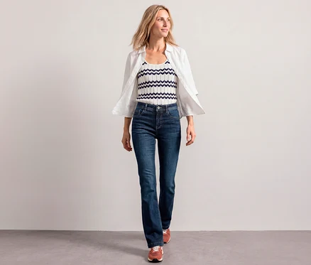 Une femme marche vers l'avant, portant une chemise blanche ouverte sur un top blanc et bleu marine à chevrons, et un Jean Bootcut Street One.
