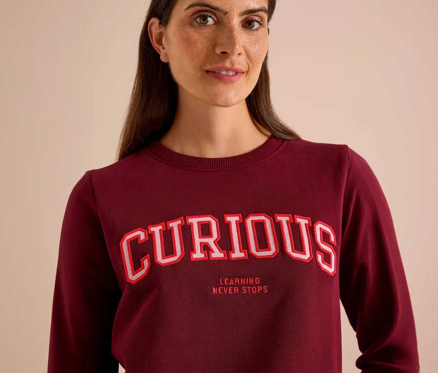Femme portant un sweatshirt CECIL bordeaux avec l'inscription « CURIOUS » et « LEARNING NEVER STOPS ».
