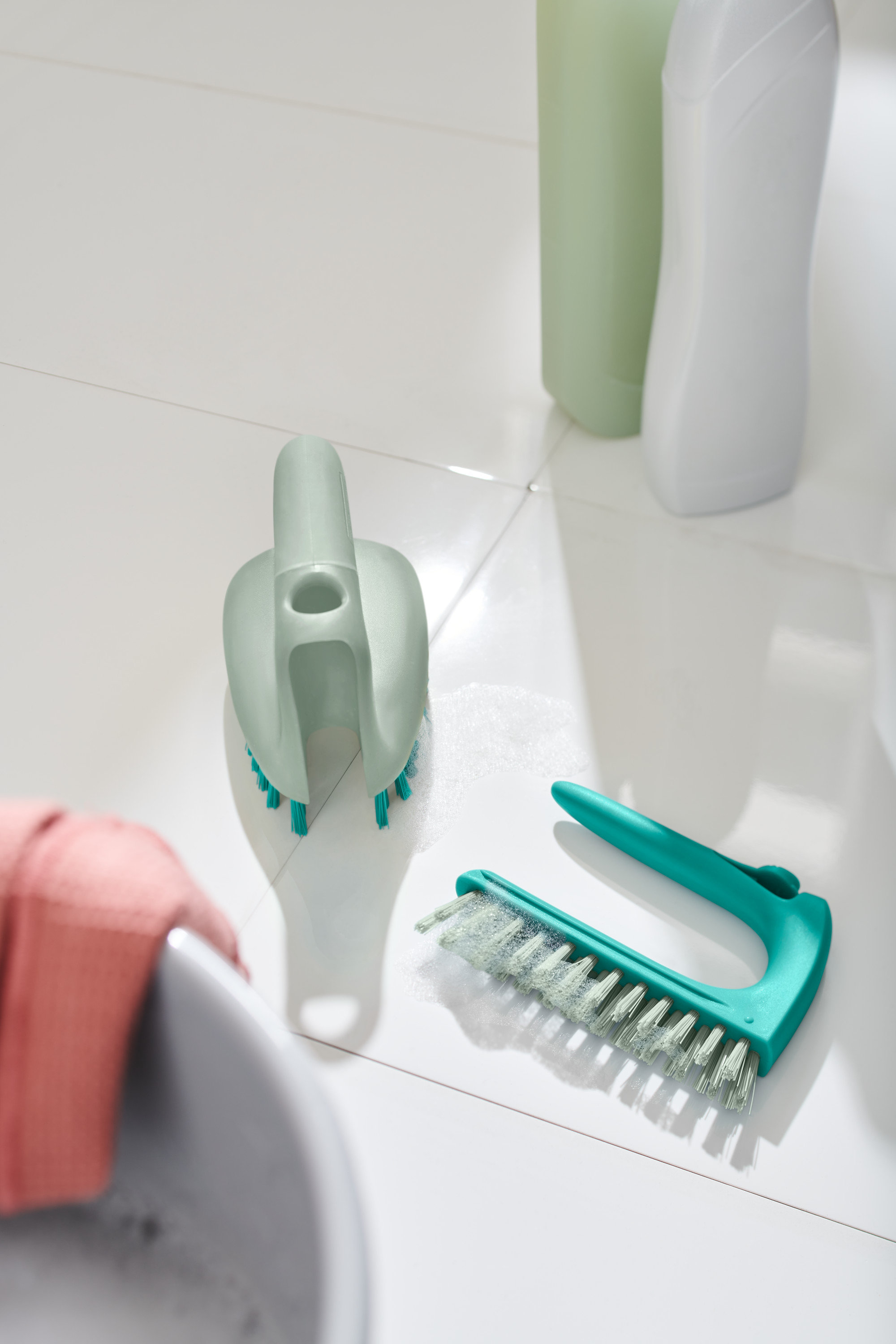 Deux brosses à sols et carrelage 2 en 1 sont posées sur des carreaux blancs à côté d'un lavabo.