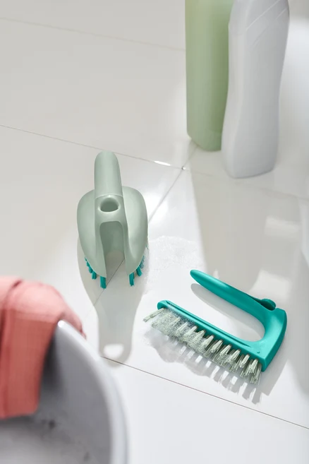 Deux brosses à sols et carrelage 2 en 1 sont posées sur des carreaux blancs à côté d'un lavabo.