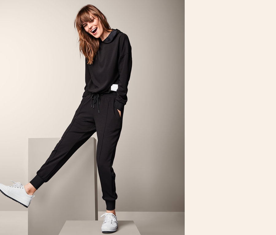 Femme posant sur des cubes gris en pantalon d’intérieur noir et sweat à capuche.