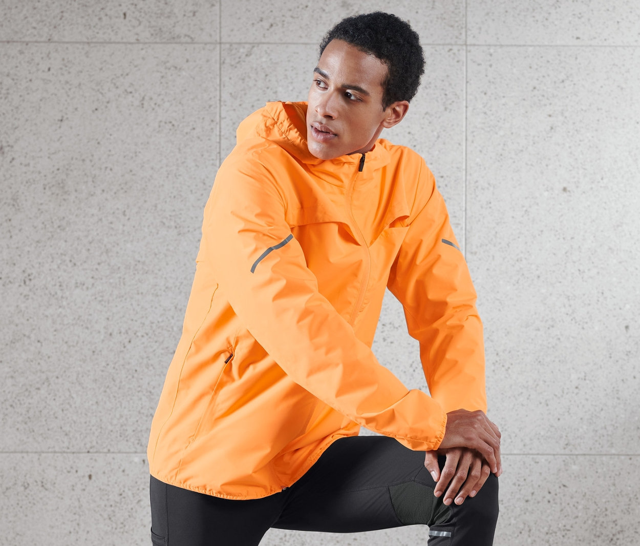 Un homme pose, une jambe sur un tabouret en bois, portant une veste coupe-vent orange fluo.