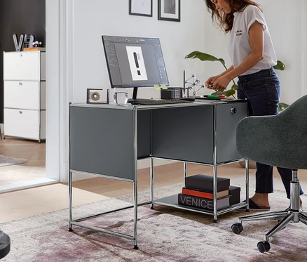 Une femme se tient à un bureau en métal « CN3 » gris avec une porte pliante et travaille.