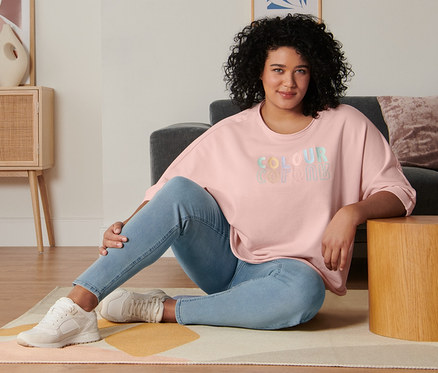 Une femme est assise sur le sol du salon et porte un sweat-shirt manches 3/4 rose, un tregging et des baskets en cuir velours.