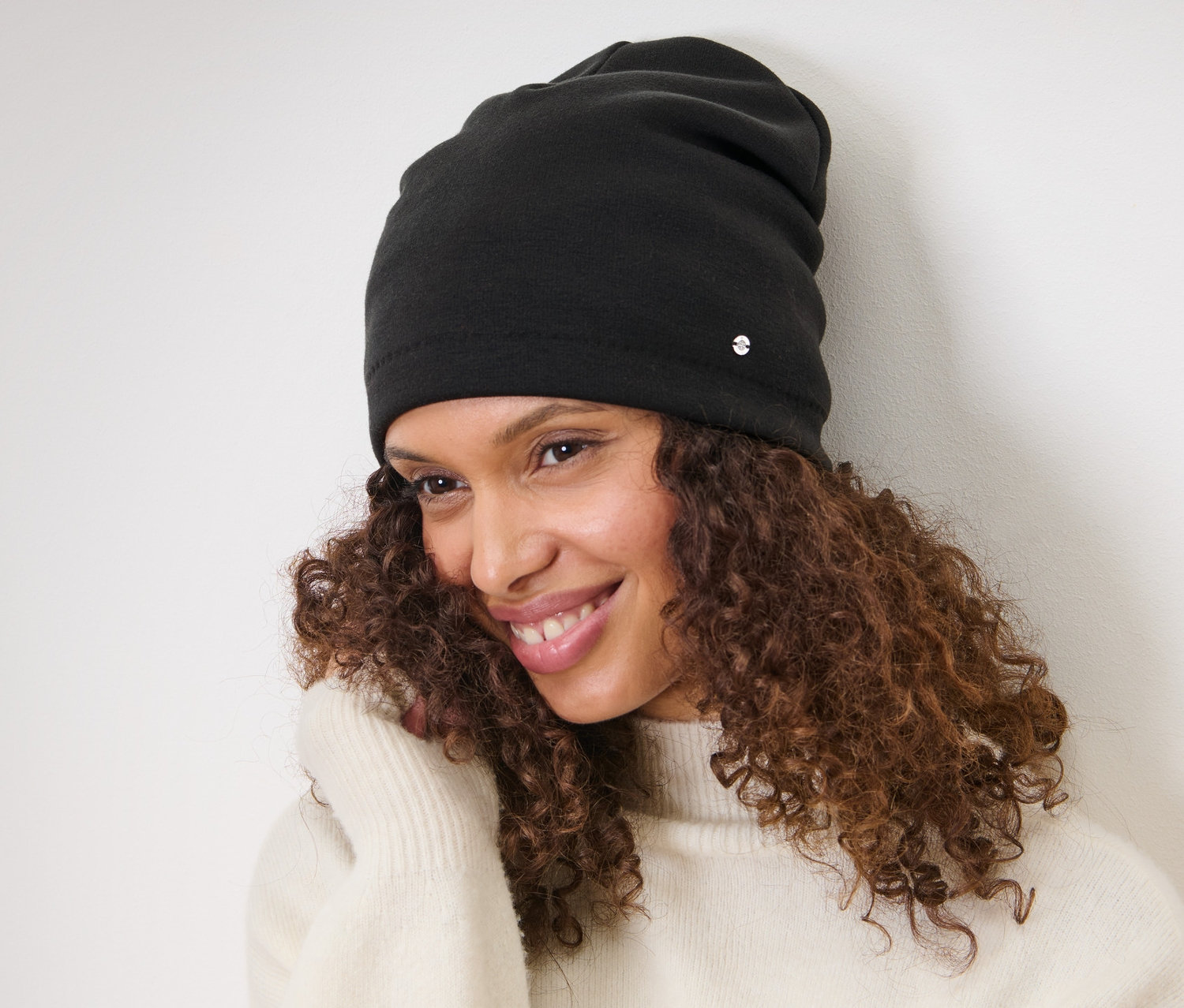 Femme aux cheveux bruns bouclés souriant et portant un bonnet en sweat noir.