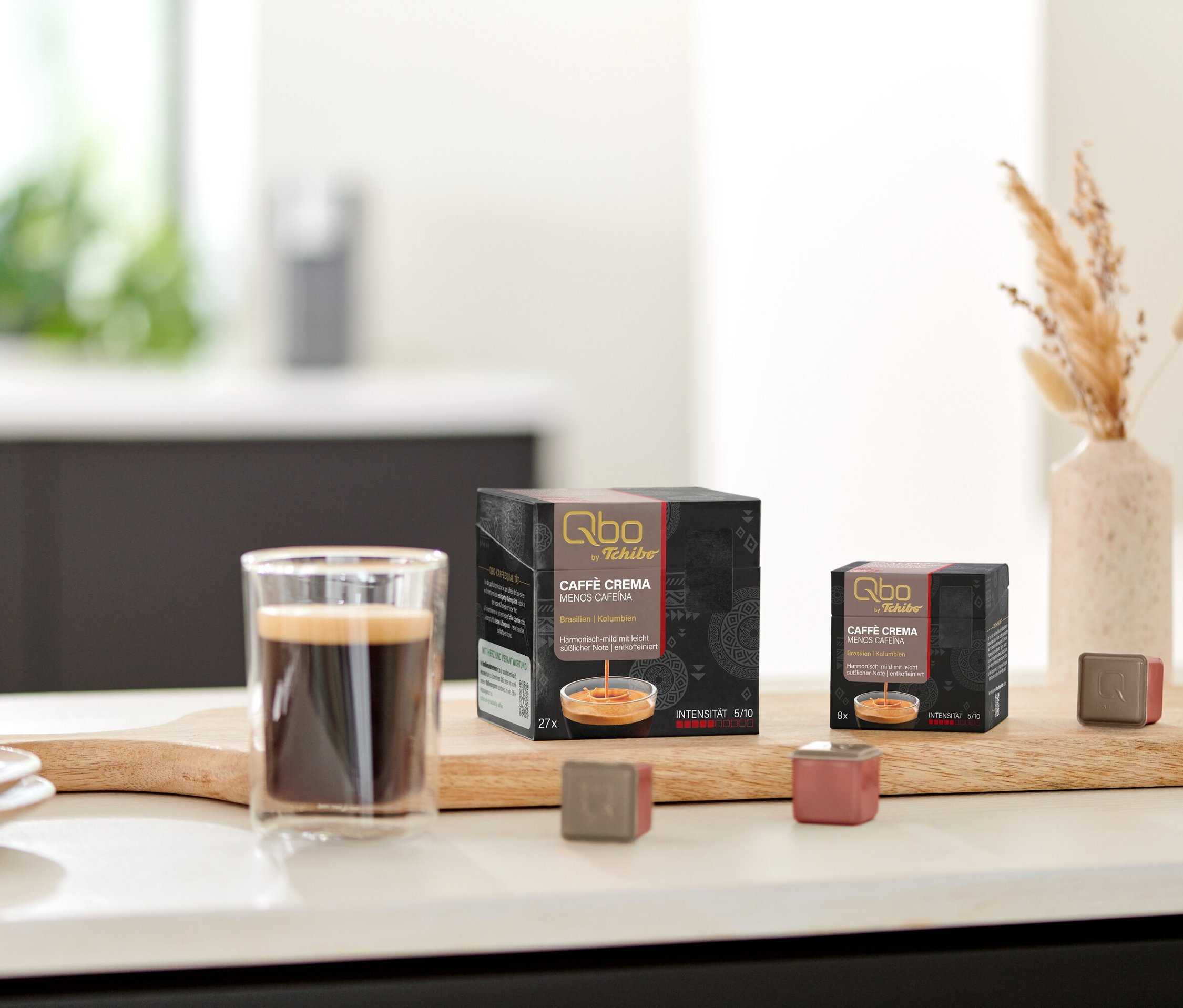 Sur une table, des paquets de capsules Qbo Caffè MENOS CAFEÍNA et une tasse de café sur une planche en bois.