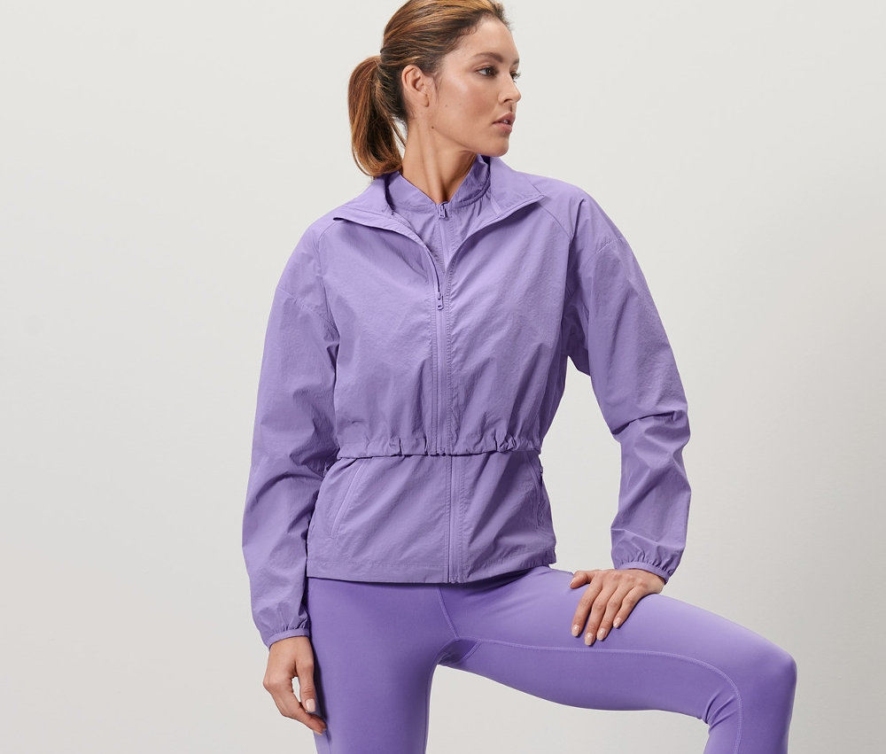 Femme posant en veste de sport 3-en-1 lilas et leggings, une jambe sur une caisse en bois.