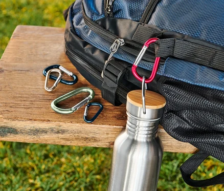 Un sac à dos bleu est posé sur un banc en bois à côté d'un jeu de 5 mousquetons et d'une bouteille d'eau en acier inoxydable.