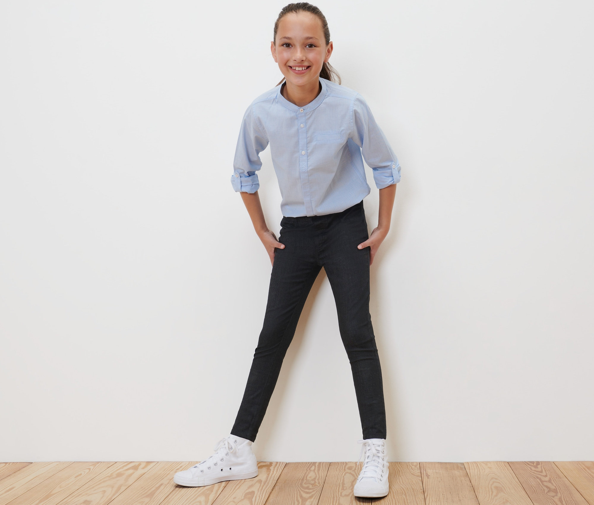 Une fille pose en tregging noir pour enfant – Fit « Jule », une chemise bleu clair et des baskets blanches.