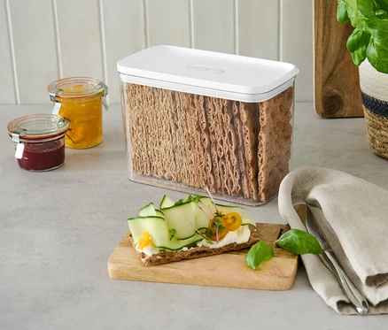 Une boîte de rangement avec un couvercle blanc remplie de crackers de pain se trouve sur un comptoir à côté de pots de confiture.