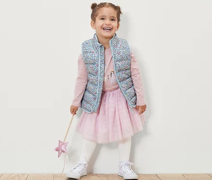 Une fillette souriante porte un gilet sans manches matelassé à fleurs, un t-shirt rose à manches longues, une jupe rose et des baskets blanches, et tient une baguette magique.