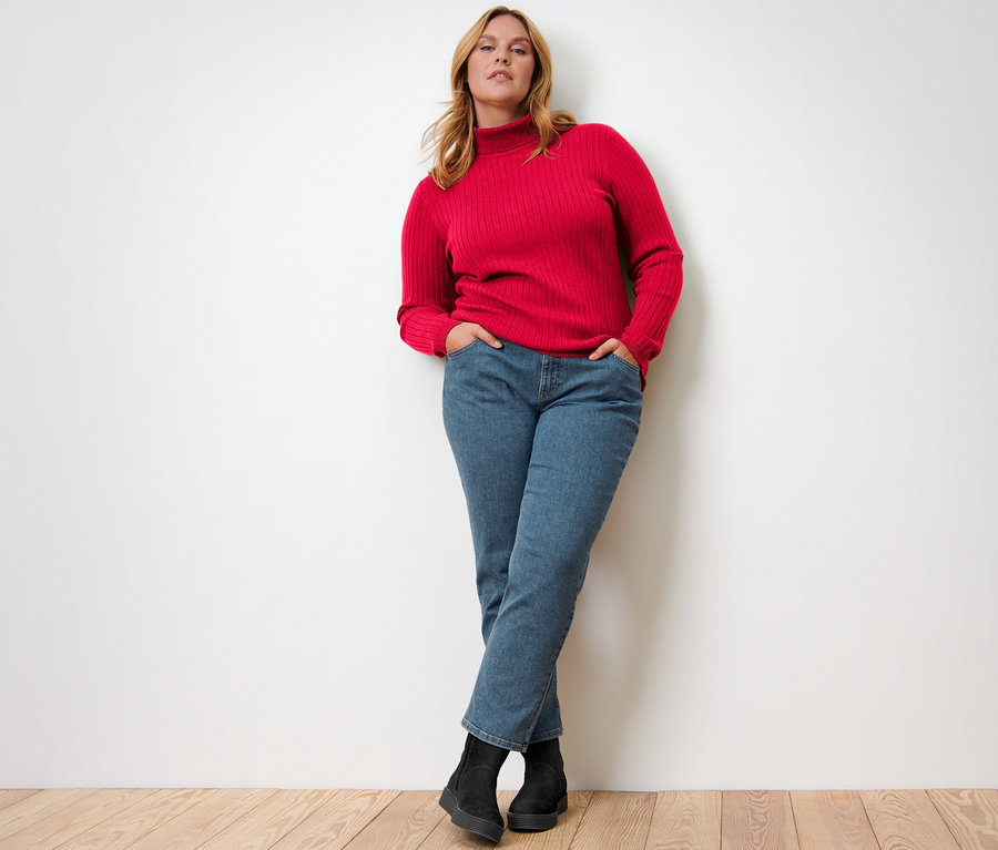 Femme posant en pull à col roulé côtelé rouge, jean et bottes noires.