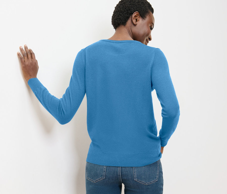 Une femme est appuyée contre un mur blanc et porte un pull en maille fine mérinos bleu clair et un jean.
