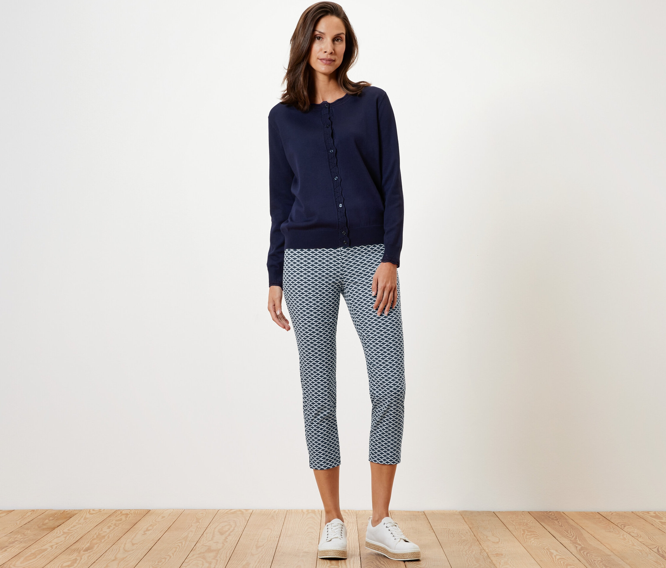 Une femme debout portant un pantalon stretch imprimé 7/8, un cardigan bleu marine et des baskets blanches.